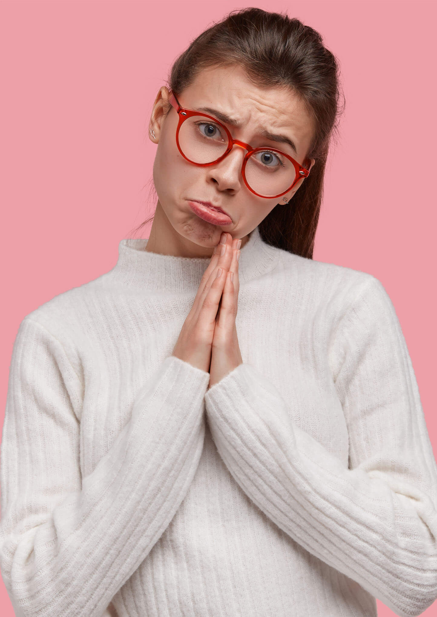 Portrait of pleading woman purses lips, keeps palms in praying gesture, asks for fortune, wears red spectacles, white casual jumper, models against pink background, has sad facial expression